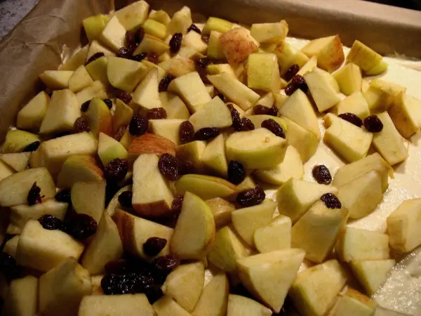 In einem Backblech liegen gehackte Äpfel und Rosinen, bereit für den Apfelkuchen mit Dinkelmehl und marmorierten Streuseln.