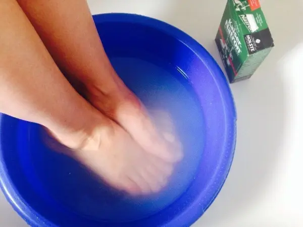 Zwei Füße sind in einem blauen Behälter mit Wasser eingetaucht, neben dem steht eine Packung Kaiser Natron für das Fußbad.
