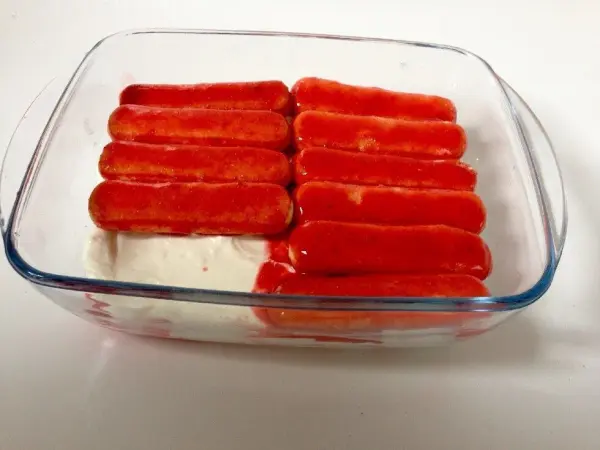 Biskuits mit Erdbeer-Glasur liegen geschichtet in einer Glasform auf einer Schicht Mascarpone-Creme in heller Umgebung.