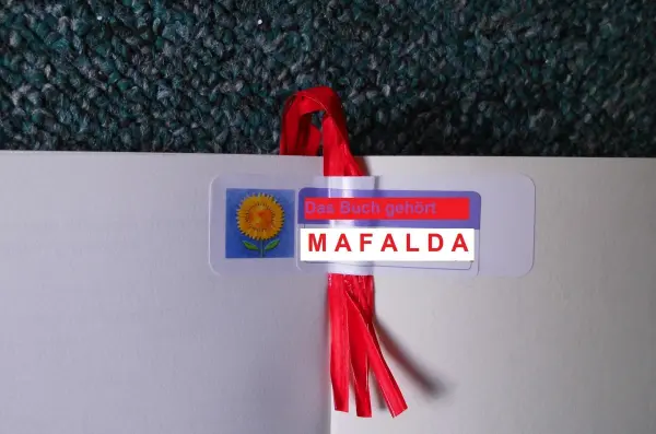 Ein Lesebändchen aus rotem Material mit einem angehängten, personalisierten Schild, das den Namen "MAFALDA" zeigt.