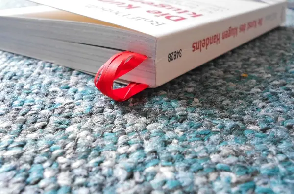 Ein rotes Lesebändchen steckt in einem offenen Buch, das auf einem bunten Teppich liegt, um das Lesen zu erleichtern.