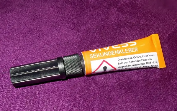 Ein Tubenbehälter mit Sekundenkleber liegt auf einem lila Stoff, bereit zum Kleben. Gefährliche Chemikalien sind gekennzeichnet.
