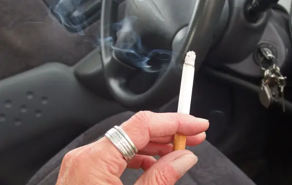 Eine Hand hält eine Zigarette, während Rauch im Auto aufsteigt, was den typischen Rauchgeruch im Innenraum verstärken kann.