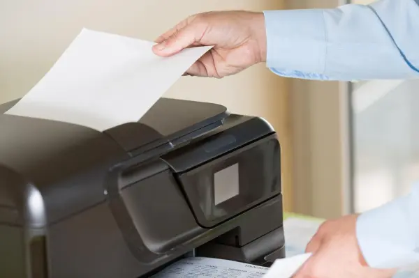 Eine Hand legt ein Blatt Papier in einen Drucker, während der Drucker in einem hellen Raum steht.