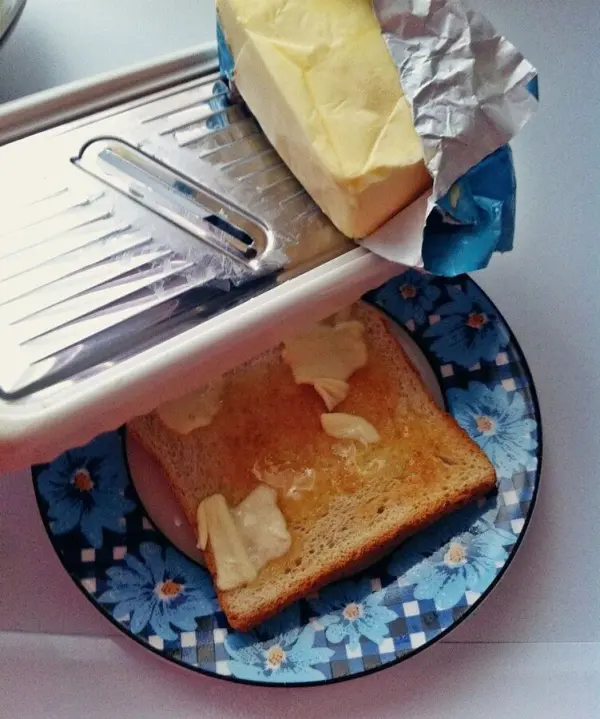 Ein Stück trockener Toast liegt auf einem bunten Teller, während ein Stück harte Butter aus der Verpackung darauf verteilt wird.