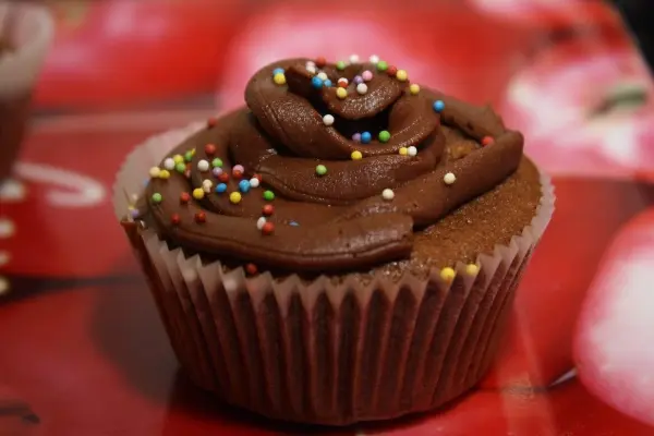 Ein Muffin mit Schokoladencreme und bunten Streuseln sitzt auf einem dekorativen Hintergrund mit roten Motiven.