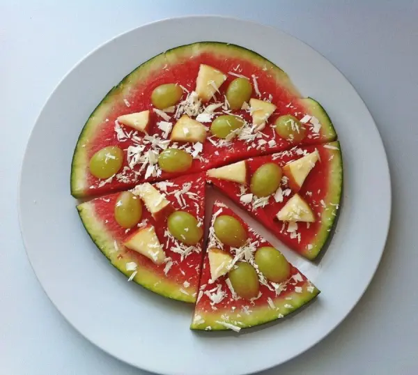 Eine Melonenscheibe, belegt mit Trauben, Ananasstücken und geraspelter weißer Schokolade, liegt auf einem Teller.