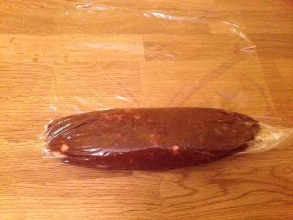 Eine Schokoladenwurst liegt in transparenter Folie auf einem Holzuntergrund. Sie wirkt fest und hat eine dunkle, schokoladige Farbe.