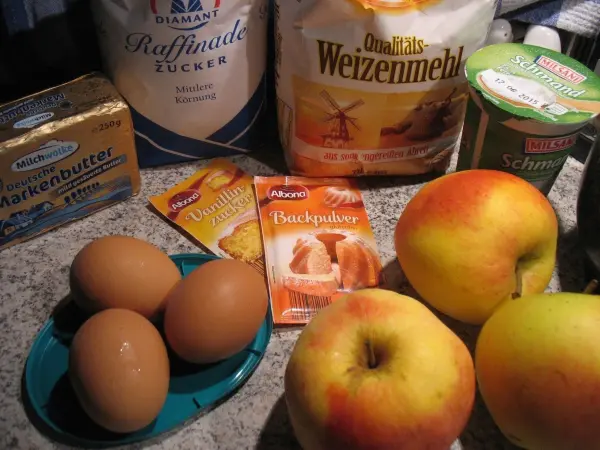 Vier Eier liegen auf einem Teller, umgeben von Äpfeln und Backzutaten wie Mehl und Streuseln, bereit zum Backen.