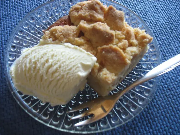 Ein Stück Apfelkuchen mit Butterstreuseln liegt auf einem Glasboden, daneben eine Portion Eiscreme auf einem blauen Tuch.
