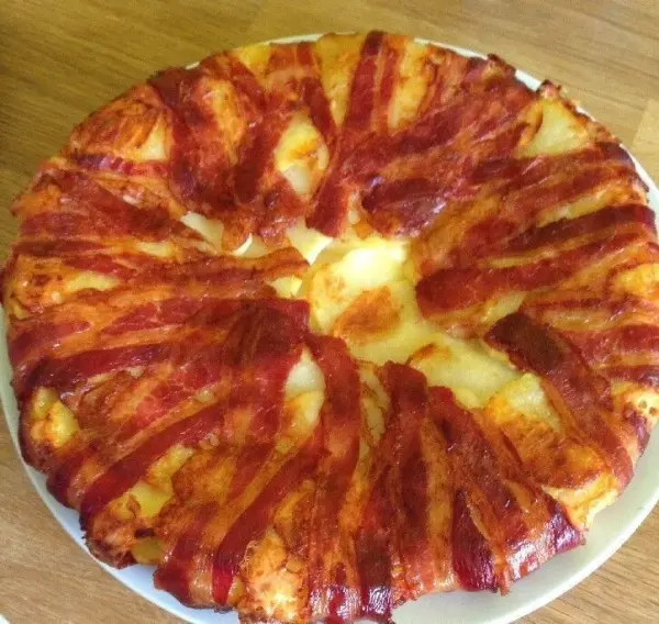 Ein runder Kartoffel-Bacon-Kuchen liegt auf einem Holzbrett und zeigt knusprigen Bacon, der attraktiv angeordnet ist.
