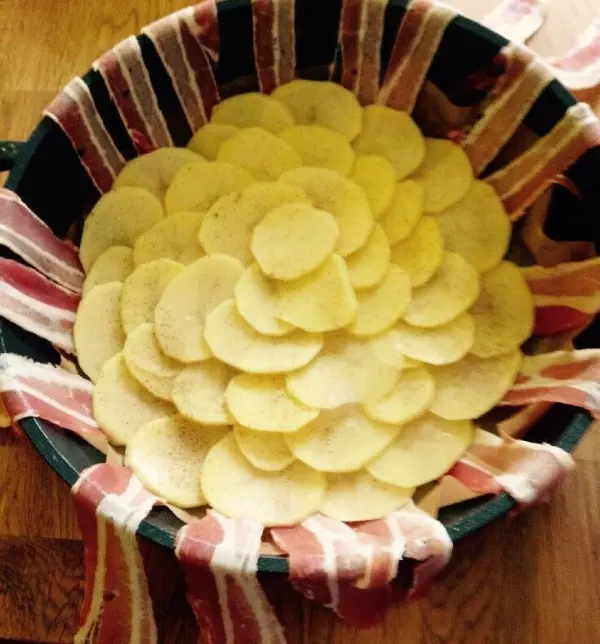 Scheiben aus Kartoffeln sind in einem Korb geschichtet, der mit Streifen von Bacon umwickelt ist, auf einer Holzoberfläche.