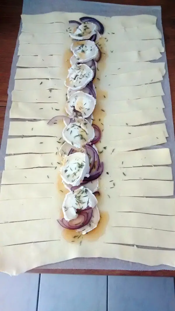 Ein Zopf aus Blätterteig liegt auf Backpapier, gefüllt mit Ziegenkäse, roten Zwiebeln und frischem Thymian.