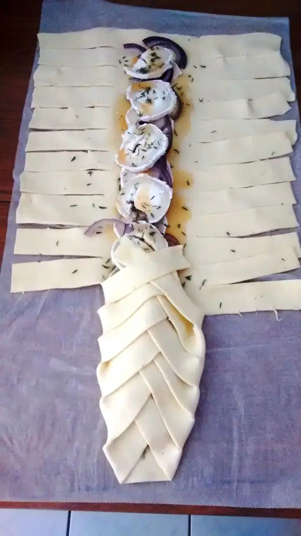 Ein Zopf aus Teig wird geflochten und mit Ziegenkäse und Thymian belegt, auf einem Backpapier auf einem Tisch.
