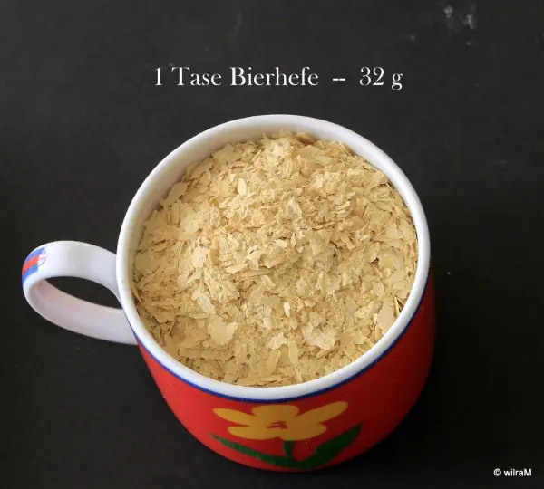 Eine rot-blumige Tasse ist gefüllt mit Bierhefe, daneben steht „1 Tasse Bierhefe = 32 g“ auf dunklem Hintergrund.