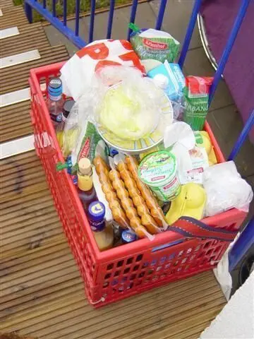 Ein roter Korb steht voll mit Lebensmitteln und Reinigungsartikeln, während um ihn herum Holzplanken zu sehen sind.