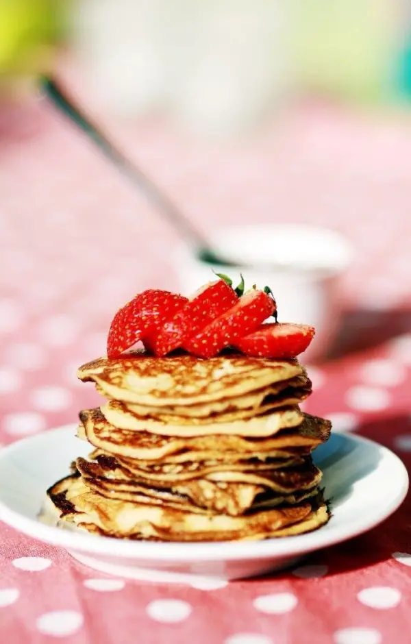 Auf einem Teller befinden sich stapelweise goldbraune Bananen-Pancakes, dekoriert mit frischen Erdbeeren. Im Hintergrund ist eine rosa, gepunktete Tischdecke zu sehen.