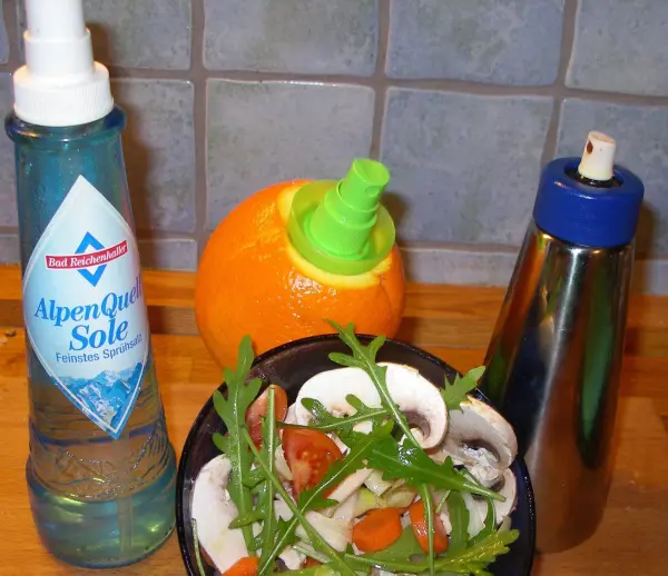 Ein Sprühbehälter für Salz und ein weiterer für Gewürze stehen neben einer Schüssel mit frischem Salat und einer Orange.