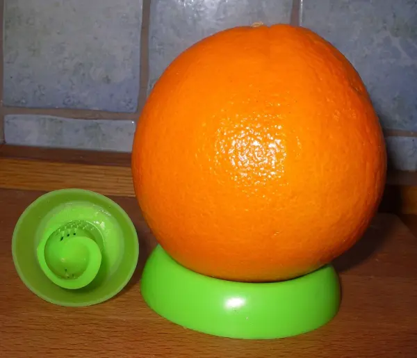 Eine orangefarbene Zitrusfrucht steht auf einem grünen Halter, während ein Sprühgerät daneben bereitsteht, um Gewürze zu sprühen.