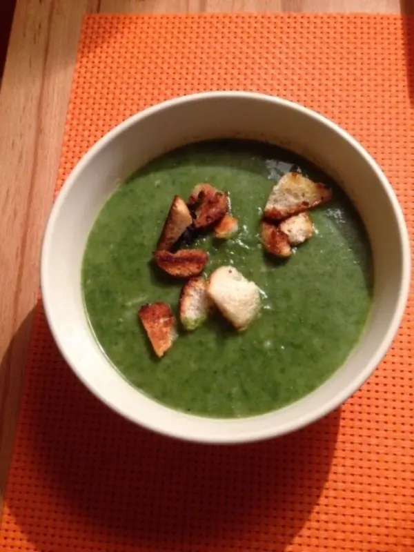 Eine grüne Spinatsuppe mit knusprigen Knoblauchcroutons in einer Schüssel, auf einem orangefarbenen Tischset.