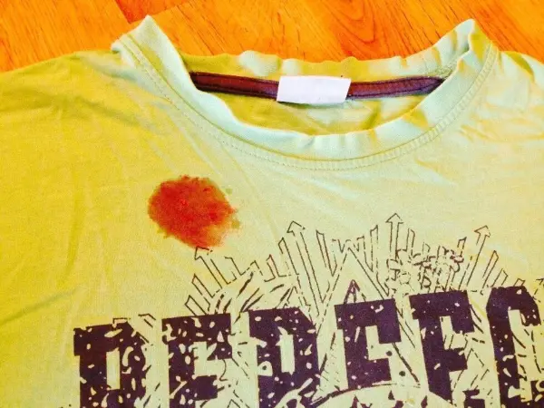 Ein T-Shirt liegt auf einem Holzfußboden, sichtbar mit einem Himbeerfleck. Der Fleck ist rot und rund, auf dem Stoff.