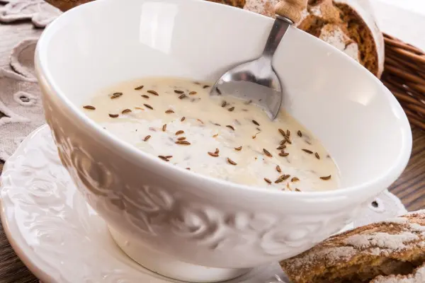 In einer dekorativen Schüssel liegt cremiger Kochkäse, garniert mit Kümmel, daneben frisches Brot im Hintergrund.