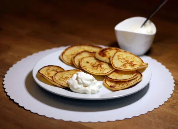 Auf einem weißen Teller liegen goldbraune Mini-Buttermilch-Pfannkuchen, daneben ein Schälchen mit Sahne, auf einem Tisch.