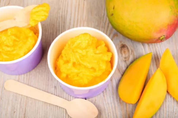 Leckeres Mangoeis wird in lila Schälchen serviert, umgeben von frischen Mango-Scheiben und einer ganzen Mango.