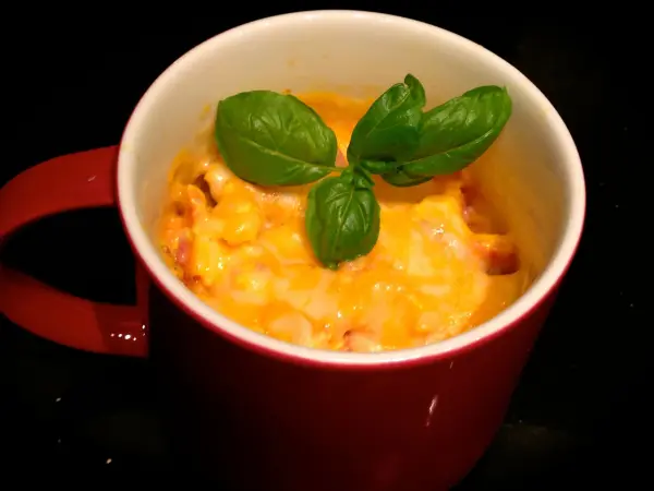 In einer roten Tasse steht Rührei, das mit Käse überbacken ist, garniert mit frischem Basilikum, auf einer dunklen Oberfläche.