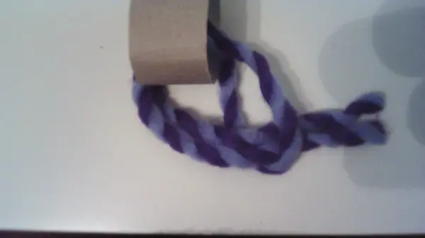 Eine lila-weiße Flaschenmütze aus Wolle wird auf einem Papprollenstück positioniert, um eine kreative Dekoidee zu präsentieren.