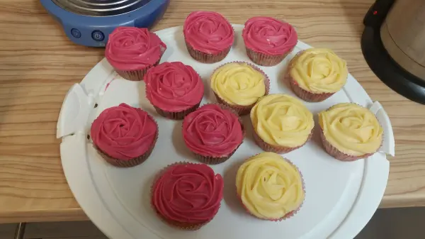 Bunte Cupcakes mit Frischkäse-Frosting in Rosa und Gelb stehen auf einem weißen Teller auf einer Holzbflächen.