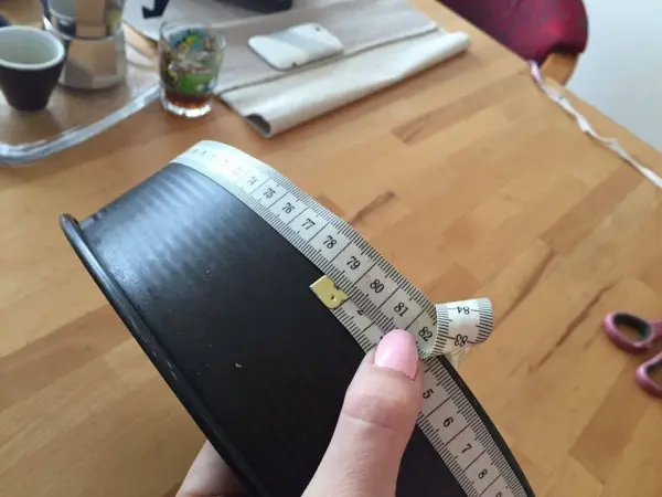 Eine Hand hält ein Maßband, das den Rand einer Kuchenform misst. Der Tisch ist mit Küchengeräten und Deko umgeben.