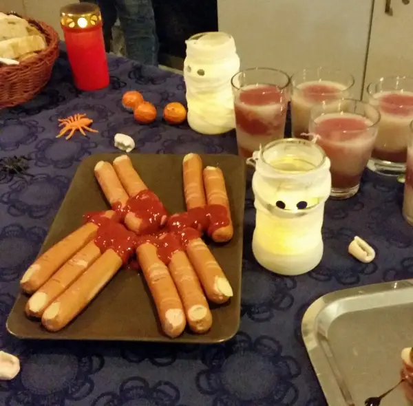 Ein Teller mit Würstchen, die mit Ketchup verziert sind, steht auf einem Tisch, umgeben von Gruselgetränken und Dekoration.
