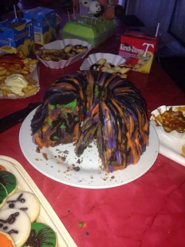 Ein bunt glasierter Kuchen in Halloween-Farben steht auf einem Teller, umgeben von Keksen und Getränken auf rotem Tisch.