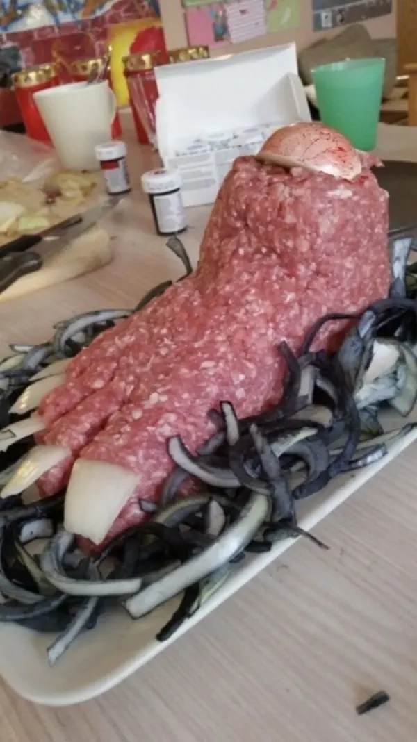Hackfleisch-Fuß ruht auf Teller, dekoriert mit geschnittenen schwarzen Zwiebeln, in einer Küche für ein Gruselbuffet.