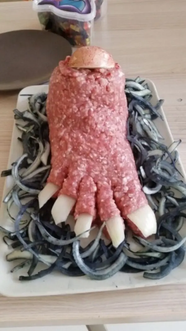 Eine lebensechte Fußform aus Hackfleisch liegt auf einer Platte, umgeben von zarten Zwiebelringen, ideal für ein Gruselbuffet.