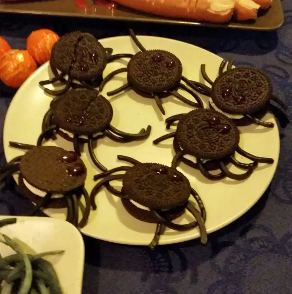 Sechs schaurige Spinnenkekse liegen auf einem Teller, umgeben von weiteren Snacks auf einem dunklen Tisch.