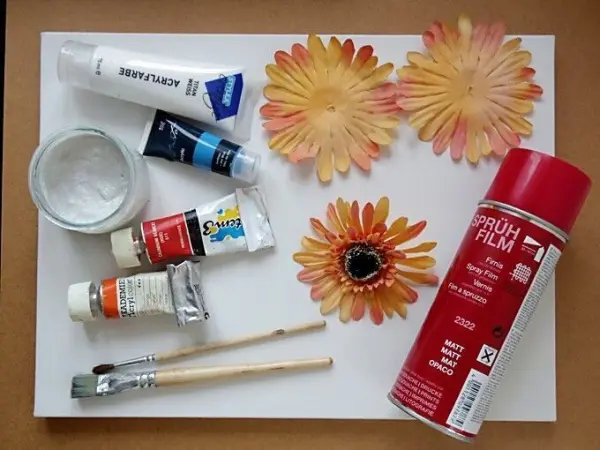 Ein Acrylbild wird mit Blütenblättern einer Plastikblume gestaltet, um kreative Kunst zu schaffen. Farbtuben und Pinsel liegen bereit.