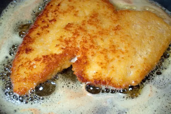 Ein goldbraun paniertes Schnitzel brät in einer Pfanne mit sprudelndem Fett, umgeben von blubbernden Blasen.