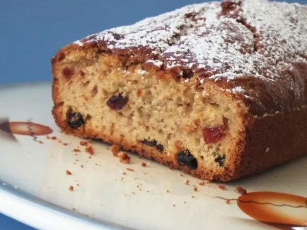Kuchenbrot mit Früchten liegt auf einem Teller, mit Puderzucker bestäubt und zeigt saftige Fruchtstücke im Inneren.