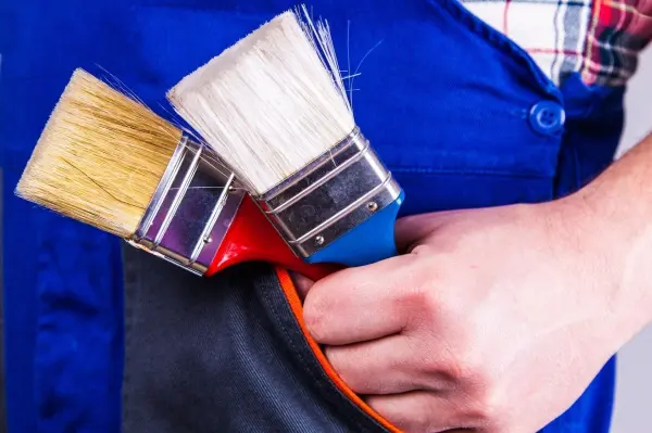 Pinsel werden von einer Hand in blauer Arbeitskleidung gehalten. Sie stecken in einer Tasche einer Jacke mit Karomuster-Hemd.
