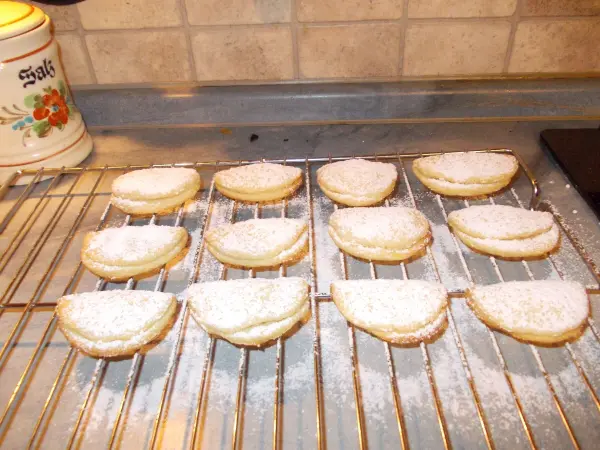 Auf einem Kuchengitter liegen gefüllte Muschelplätzchen, bestreut mit Puderzucker, in einer gemütlichen Küche.