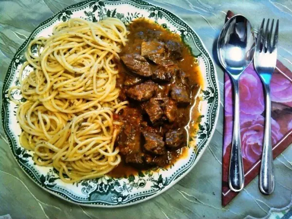 Ein Teller mit Rindergulasch und Spaghetti liegt auf einem Tisch, umgeben von Besteck und einem dekorativen Untersetzer.