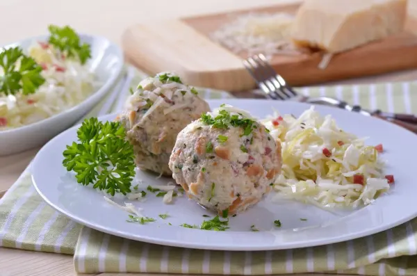 Auf einem Teller liegen zwei Tiroler Knödel, umgeben von einem frischen Salat. Im Hintergrund sind weitere Zutaten sichtbar.