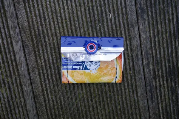 Ein Mini-Portemonnaie aus Capri-Sonne liegt auf einem holzverkleideten Untergrund, farbenfroh und gut verarbeitet.