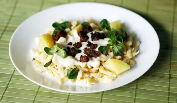 Spätzle und Kartoffeln liegen in einer Schüssel, bedeckt mit Käsesoße und frischen Kräutern, auf einem grünen Tisch.
