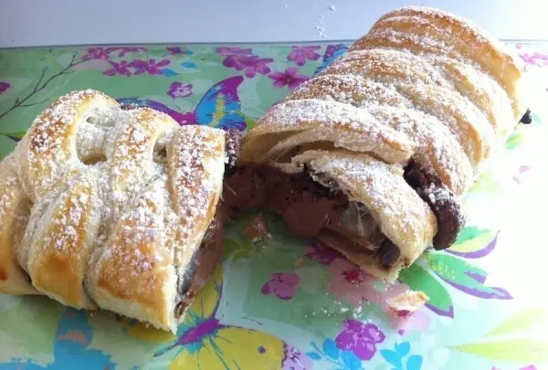 Blätterteigstrudel liegt auf einer bunten Platte, geschnitten, mit Schokolade gefüllt und mit Puderzucker bestäubt.