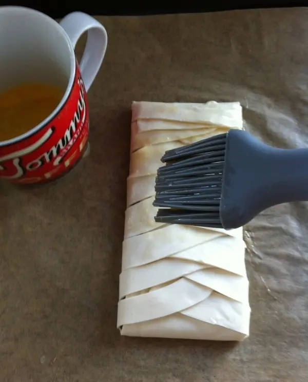 Ein Blätterteigstrudel wird mit einer Backbürste bestrichen, während eine Tasse neben ihm steht.