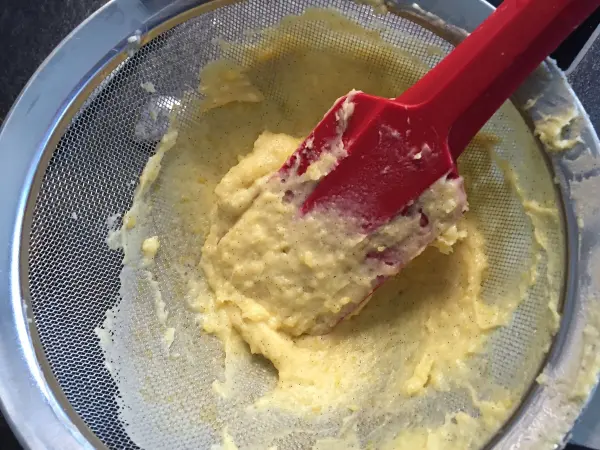Ein gelber Schokoladenteig wird mit einem roten Spatel durch ein Sieb gedrückt, um eine glatte Konsistenz zu erreichen.