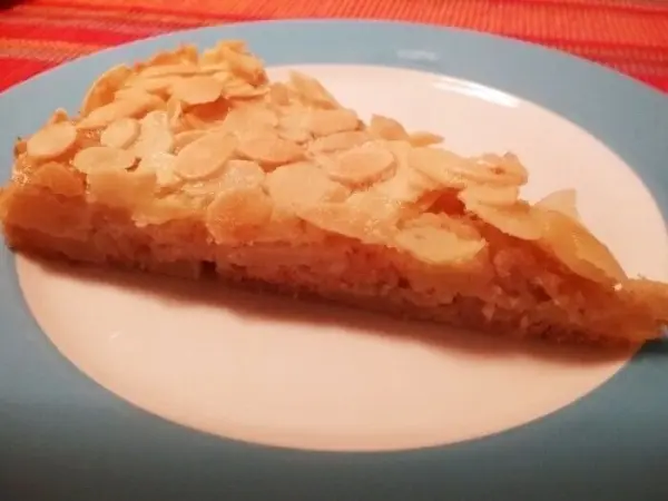 Ein Stück französischer Apfelkuchen liegt auf einem weißen Teller mit blauer Umrandung, umgeben von einer Tischdecke in warmen Farben.